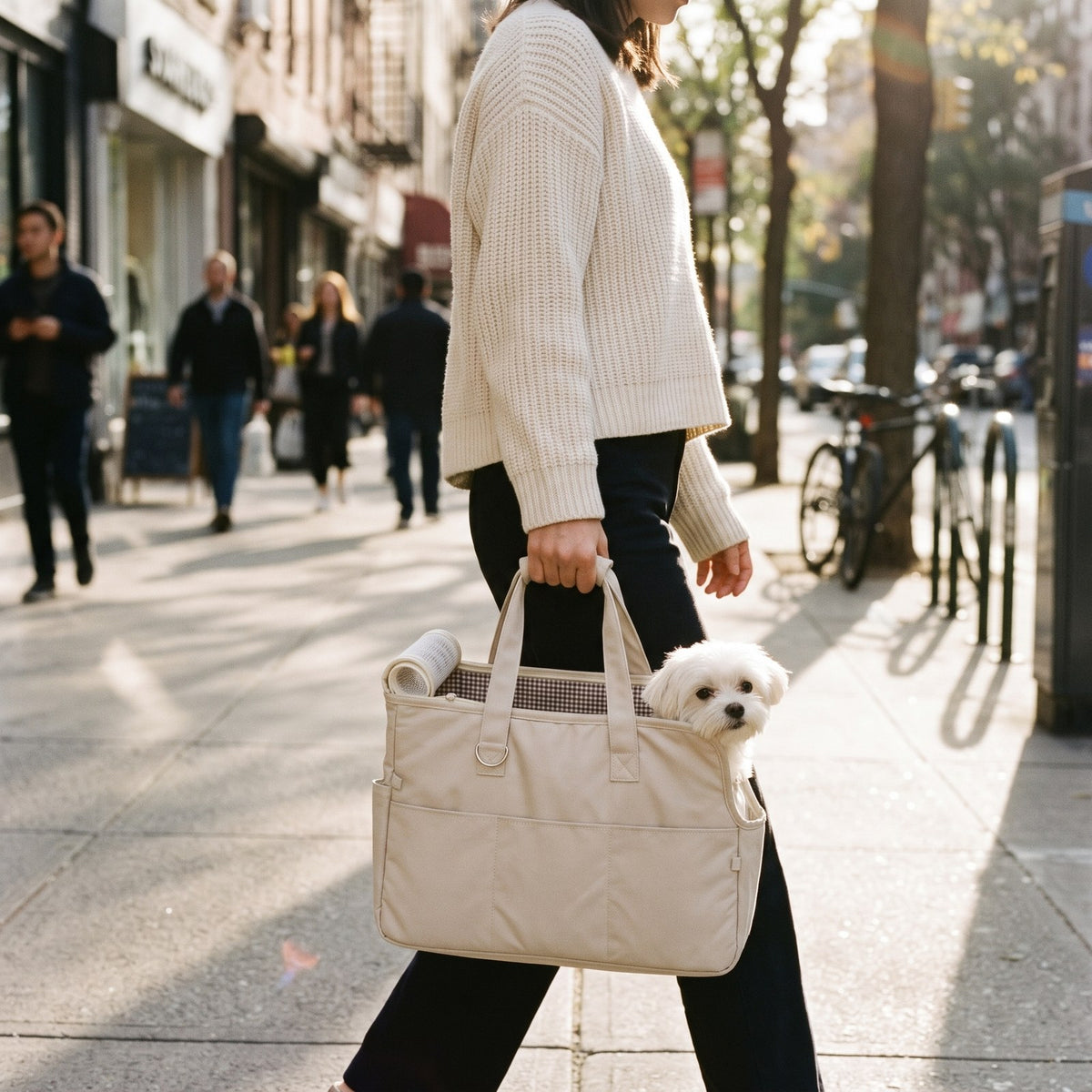 Woman carrying the beige pet travel tote on a city sidewalk with a small white dog peeking out